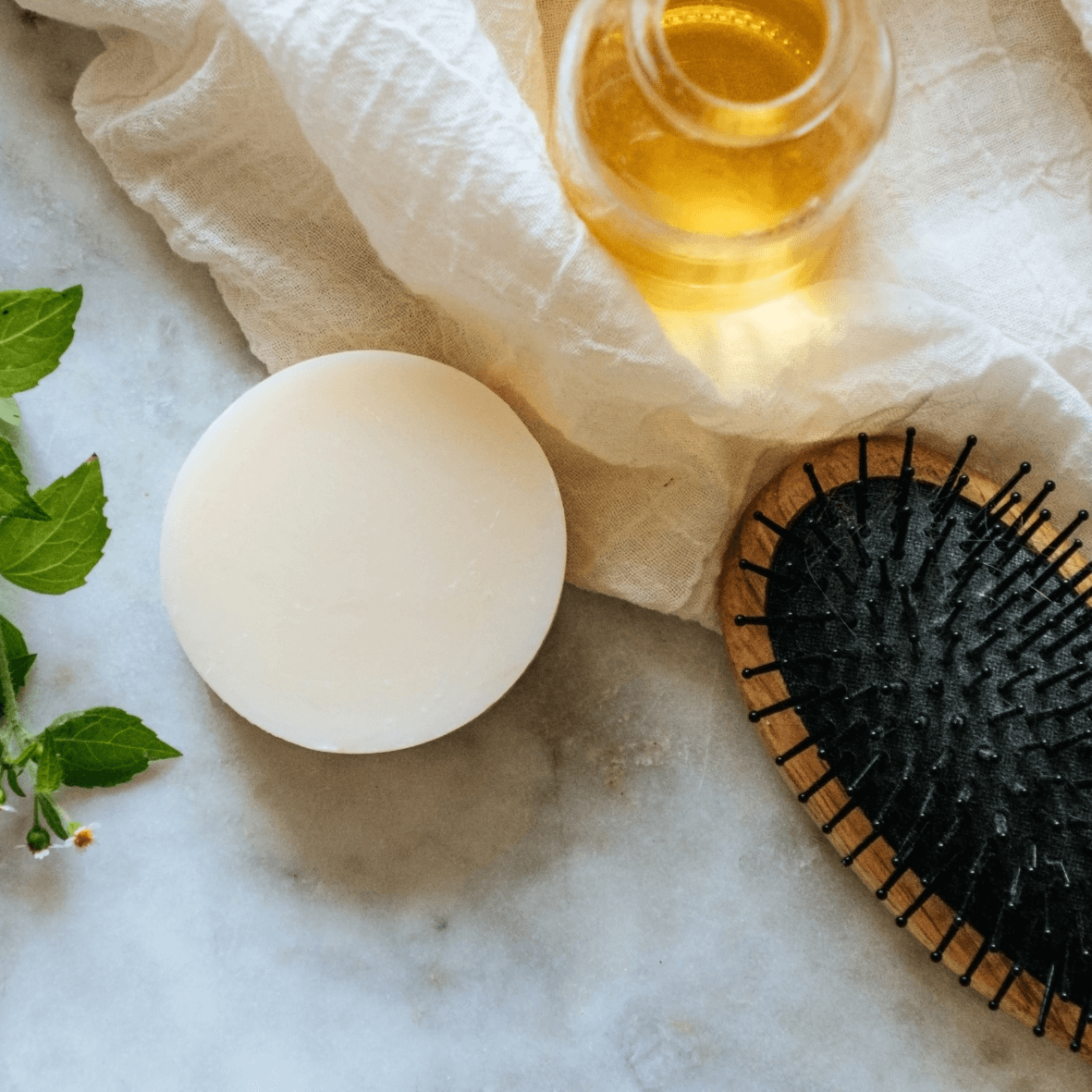 Flat lay still life with conditioner bar, hair brush, jar of oil, and mint leaves on a marble slab Alpaca Soaps AlpacaSoaps